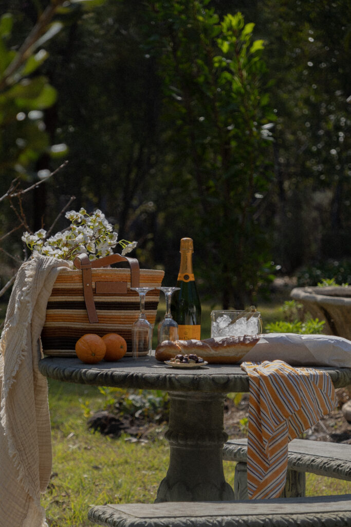 A cozy picnic setup in a backyard garden features a basket filled with fresh fruits and a bottle of sparkling wine.