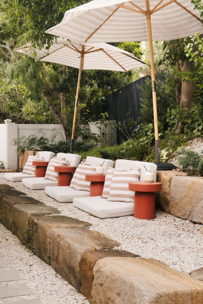 A serene retreat unfolds with cozy lounge chairs featuring striped fabric, shaded by large umbrellas, all set against a backdrop of vibrant foliage and natural stone.